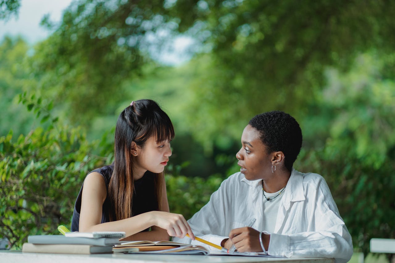 Focused multiracial female students in casual clothes sitting at table with textbooks and copybooks and discussing home assignment together while studying in lush summer garden