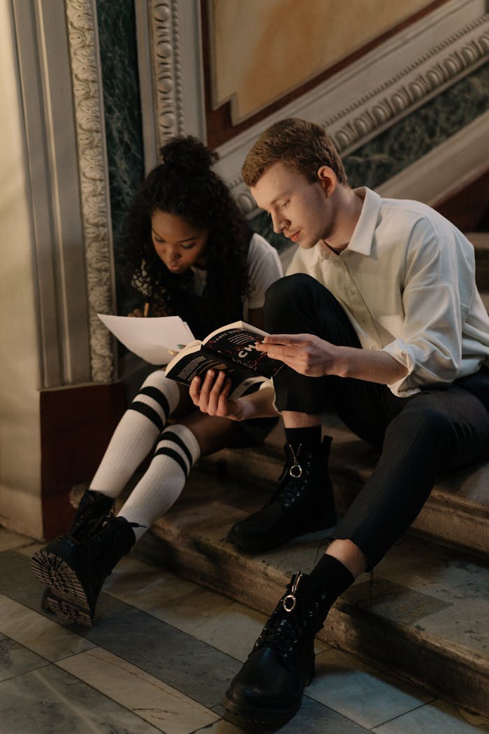 Two students sitting on staircase studying with books in university hallway.